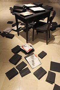 Schwarzer Tisch und Sessel, auf dem Tisch und am Boden liegen zugeklappte Bücher und offene bezeichnete Blätter