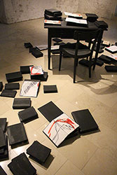 Schwarzer Tisch und Sessel, auf dem Tisch und am Boden liegen zugeklappte Bücher und offene bezeichnete Blätter